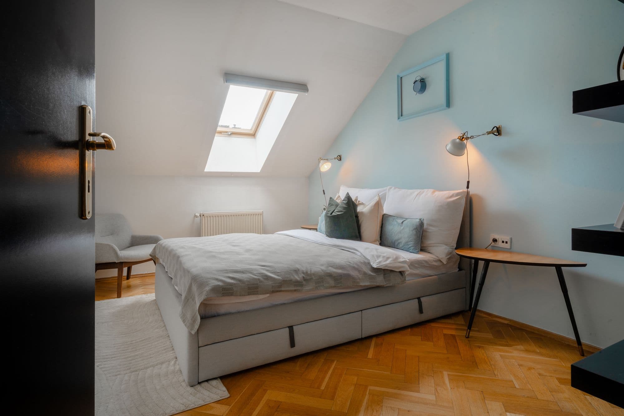 Attic bedroom with a skylight, grey upholstered bed with storage drawers, armchair and wall lamps on a blue accent wall