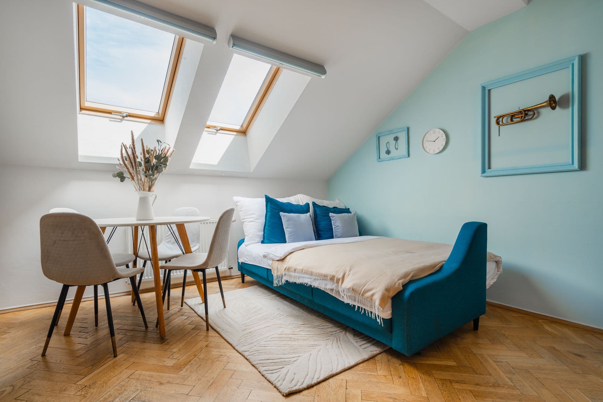 Sunny attic living room in Prague with two skylights, a teal sofa bed, round dining table and a framed trumpet on a blue wall