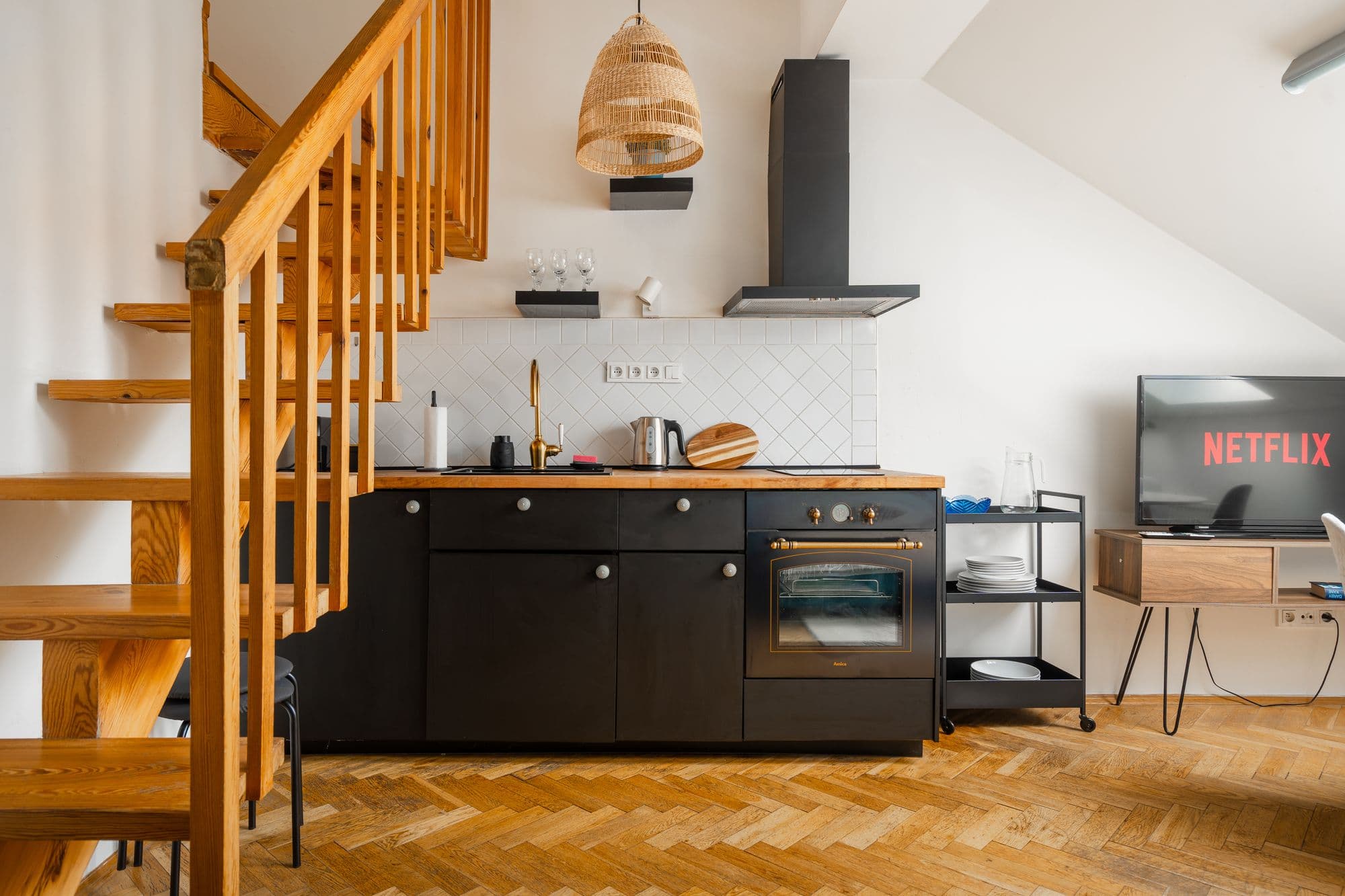 Black kitchenette with a brass faucet, retro oven and diagonal white tiles next to a wooden staircase and rattan pendant lamp