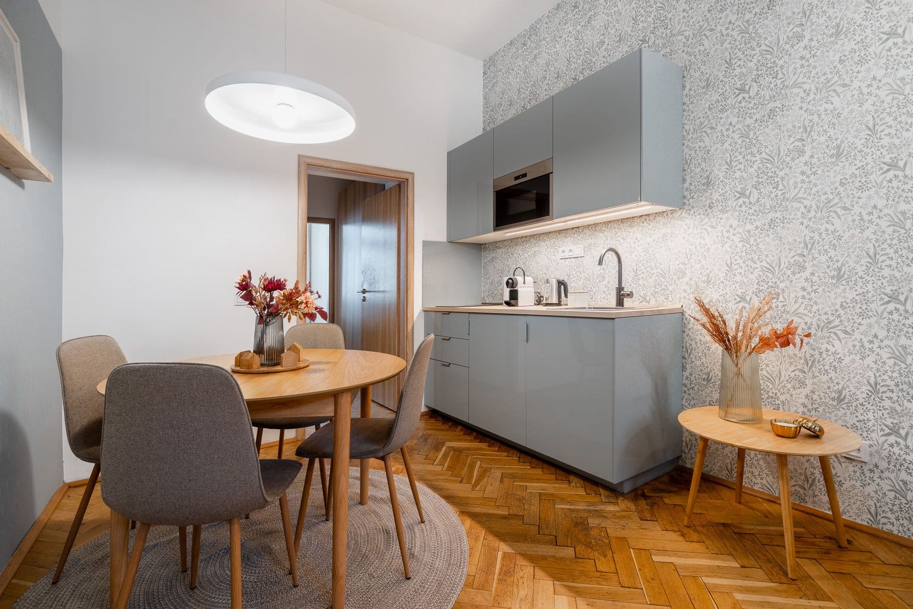 Dining nook with a round wooden table and four upholstered chairs next to a grey-blue kitchenette with botanical wallpaper