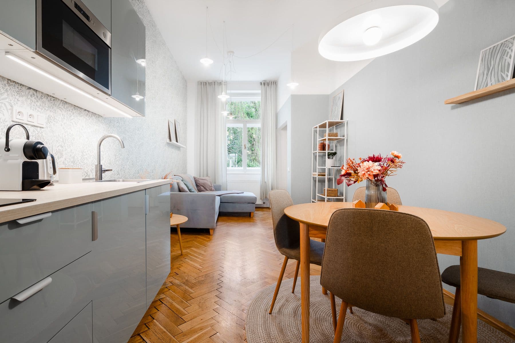 Bright living space with a grey corner sofa, tall windows onto greenery, a white shelving unit and a dining table with flowers