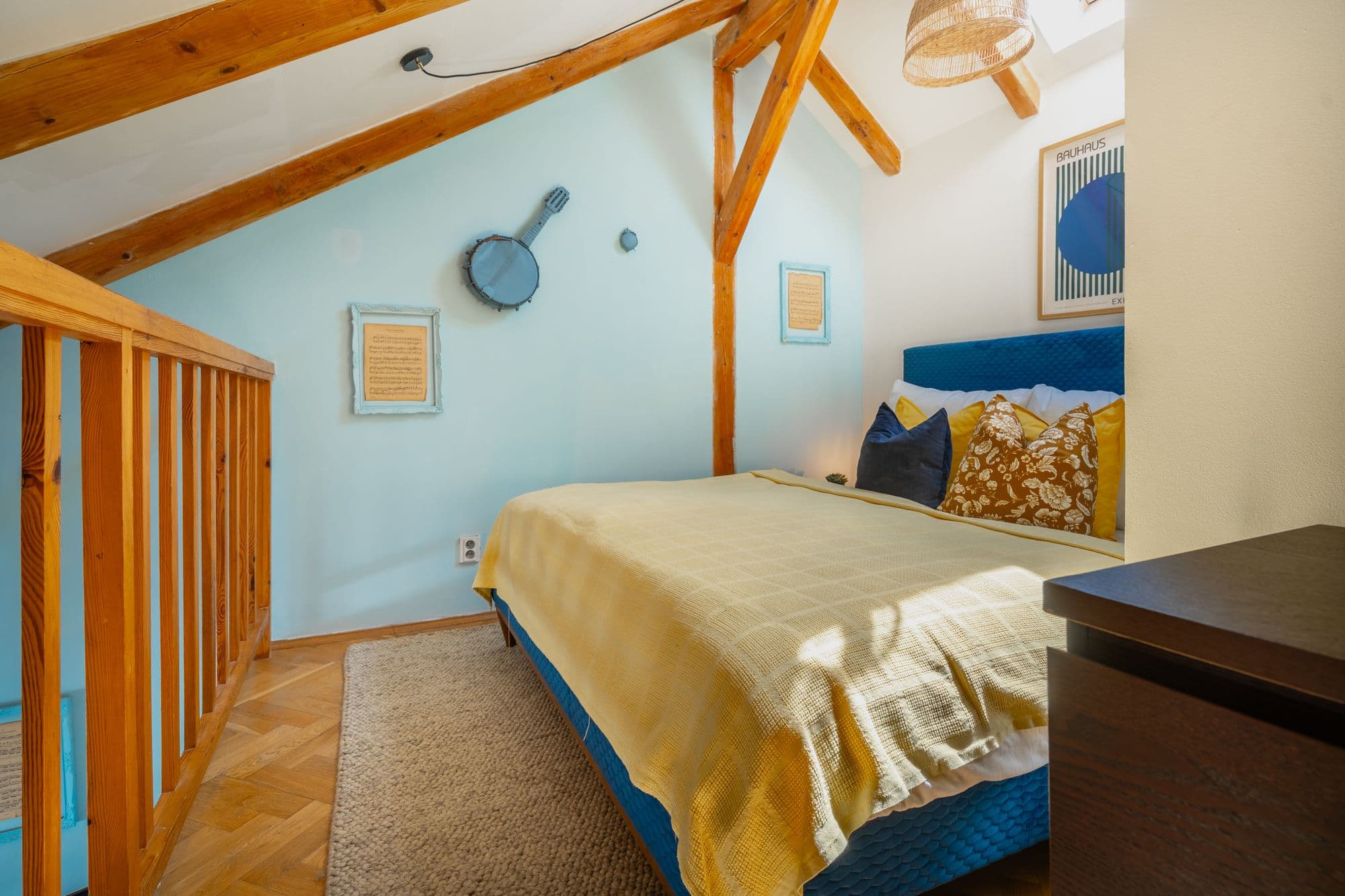 Sleeping gallery under exposed roof beams with a yellow bedspread, framed sheet music and a banjo on the wall