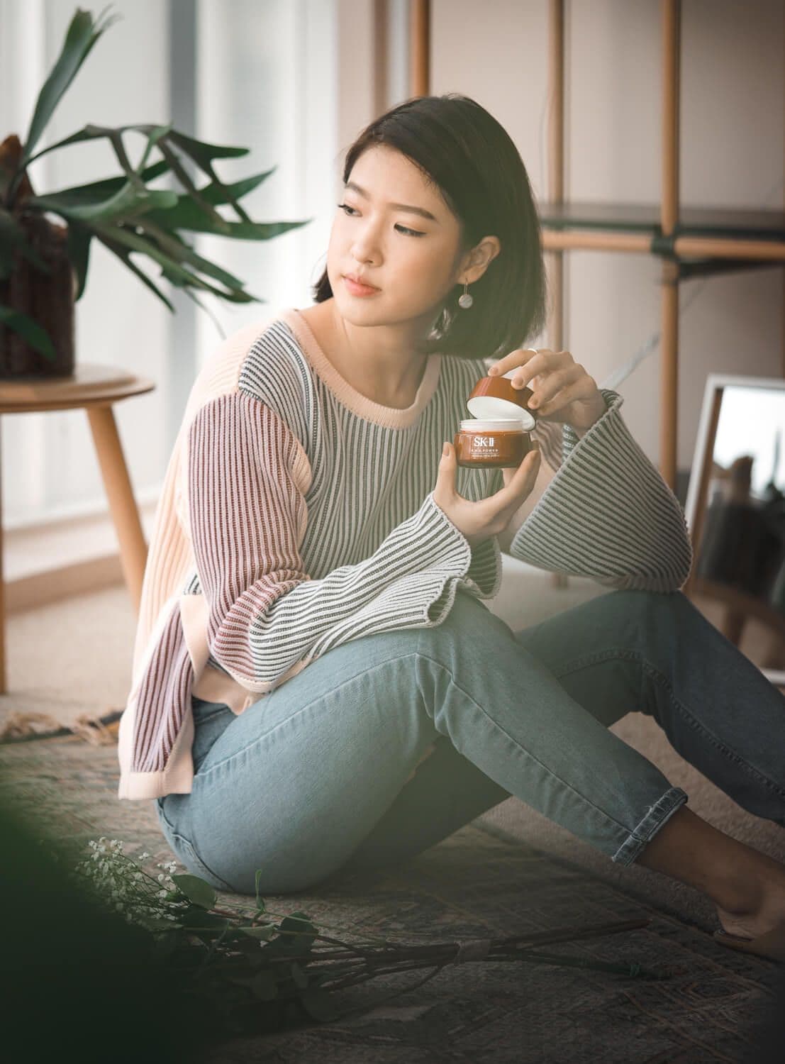 Bella Kuan sitting on a rug with an open jar of SK-II R.N.A. Power cream, looking aside.