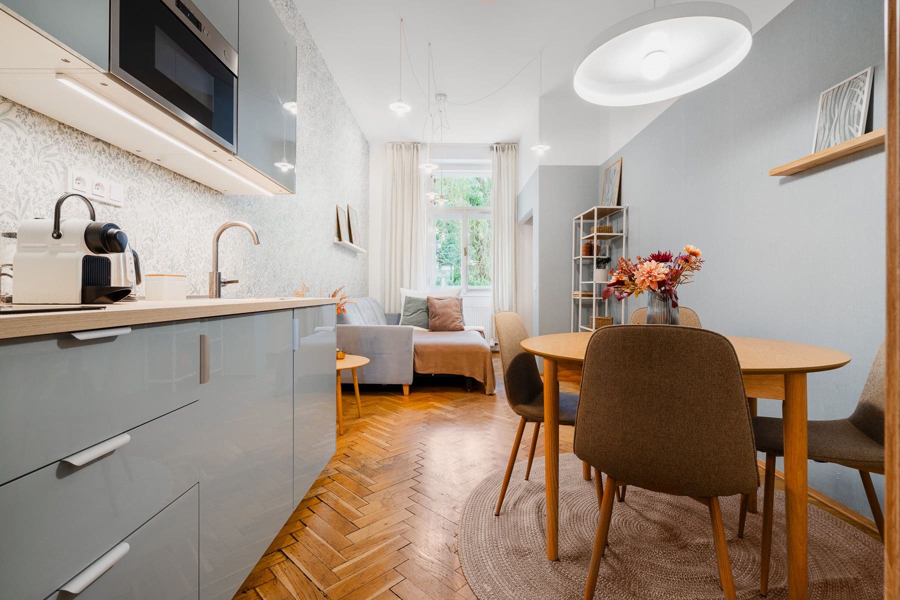 Open-plan kitchenette and dining area in a Prague apartment with herringbone parquet, a round oak table and a sofa by the window