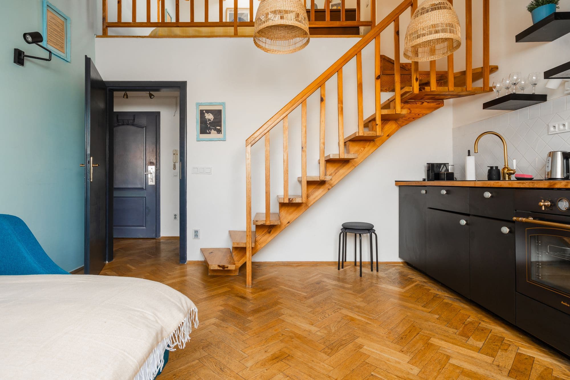 Open wooden staircase leading up to a sleeping gallery, with rattan pendant lamps and a black kitchenette below