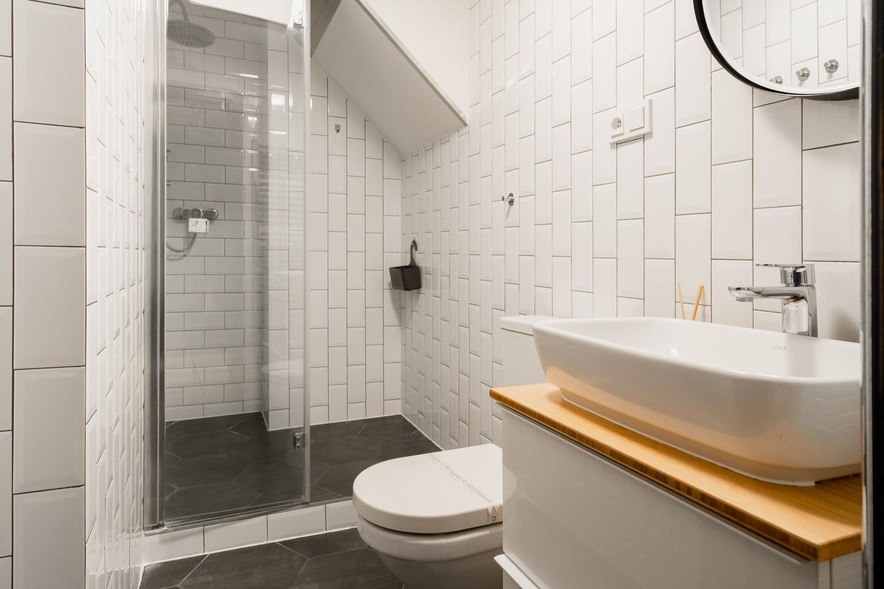 Bathroom with white subway tiles, a glass shower under the sloped ceiling, vessel sink on a bamboo counter and hexagon floor tiles
