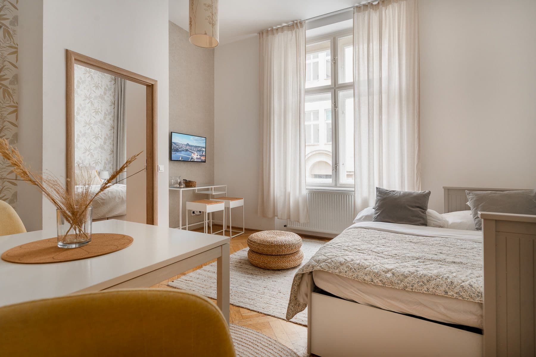 Bright living room in a Prague period apartment with a tall double window, daybed, woven poufs and a view into the bedroom