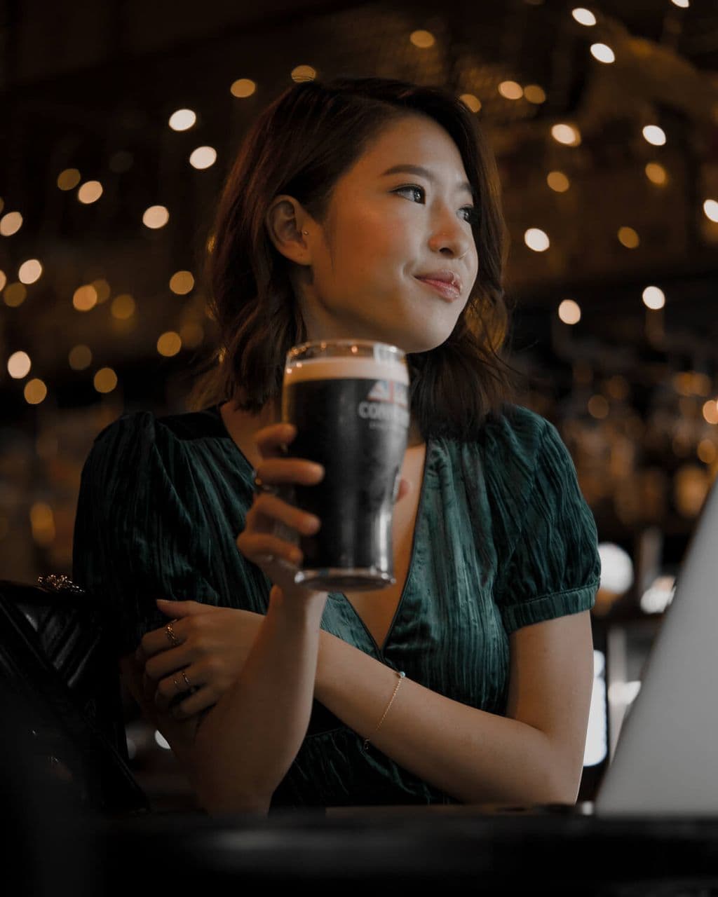 Bella Kuan in a green velvet dress holding a pint of Connor’s stout, bar lights blurred behind her.
