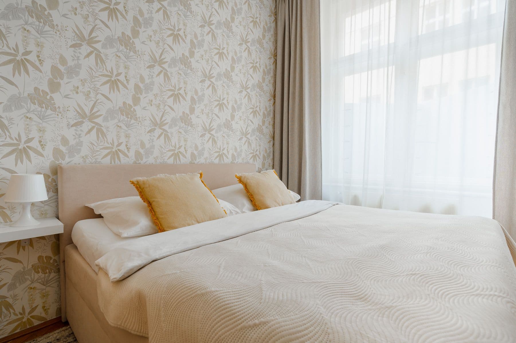 Bedroom with a beige upholstered bed, mustard fringed cushions and a textured cream bedspread beside sheer curtains