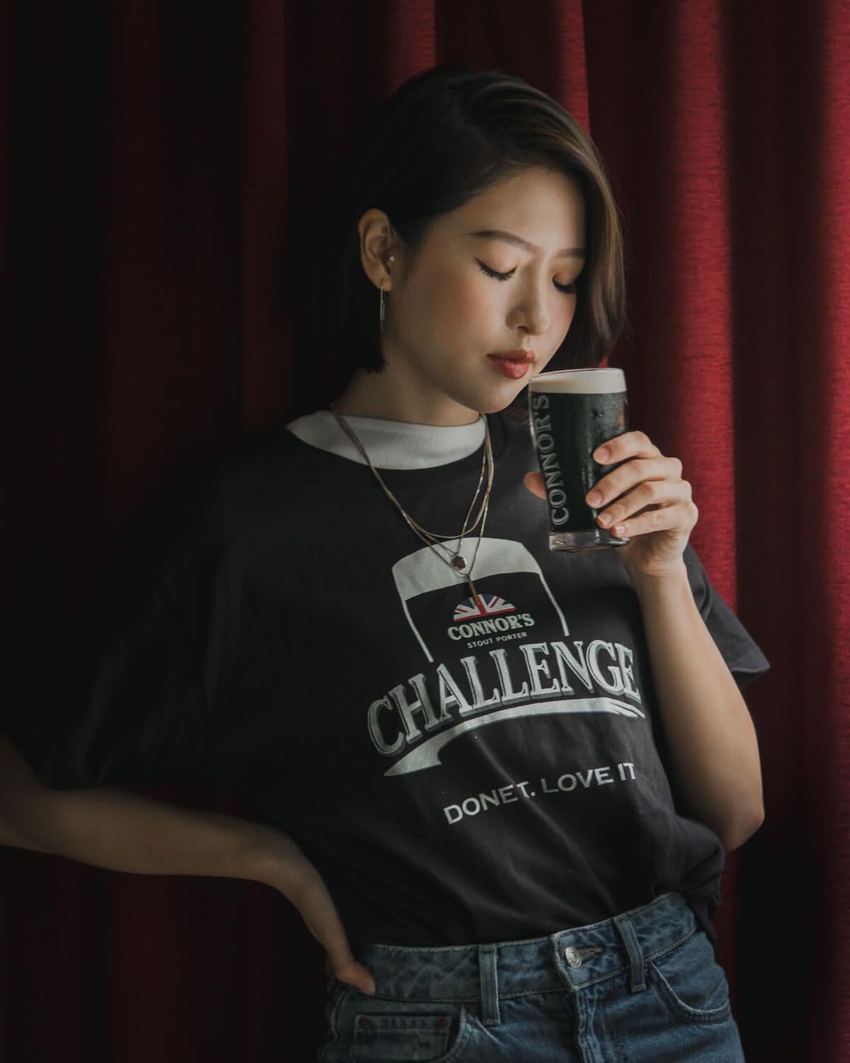 Bella Kuan in a Connor’s Stout Porter Challenge T-shirt raising a glass of stout, a red curtain behind her.