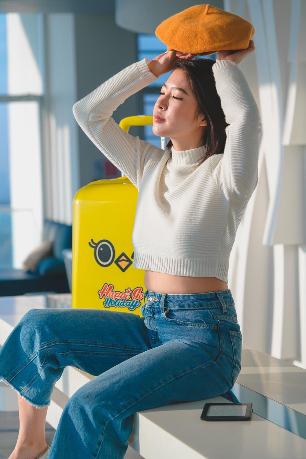 Bella Kuan in a white sweater and orange beret by a sunny window, a yellow Happi Go Holiday suitcase behind her.