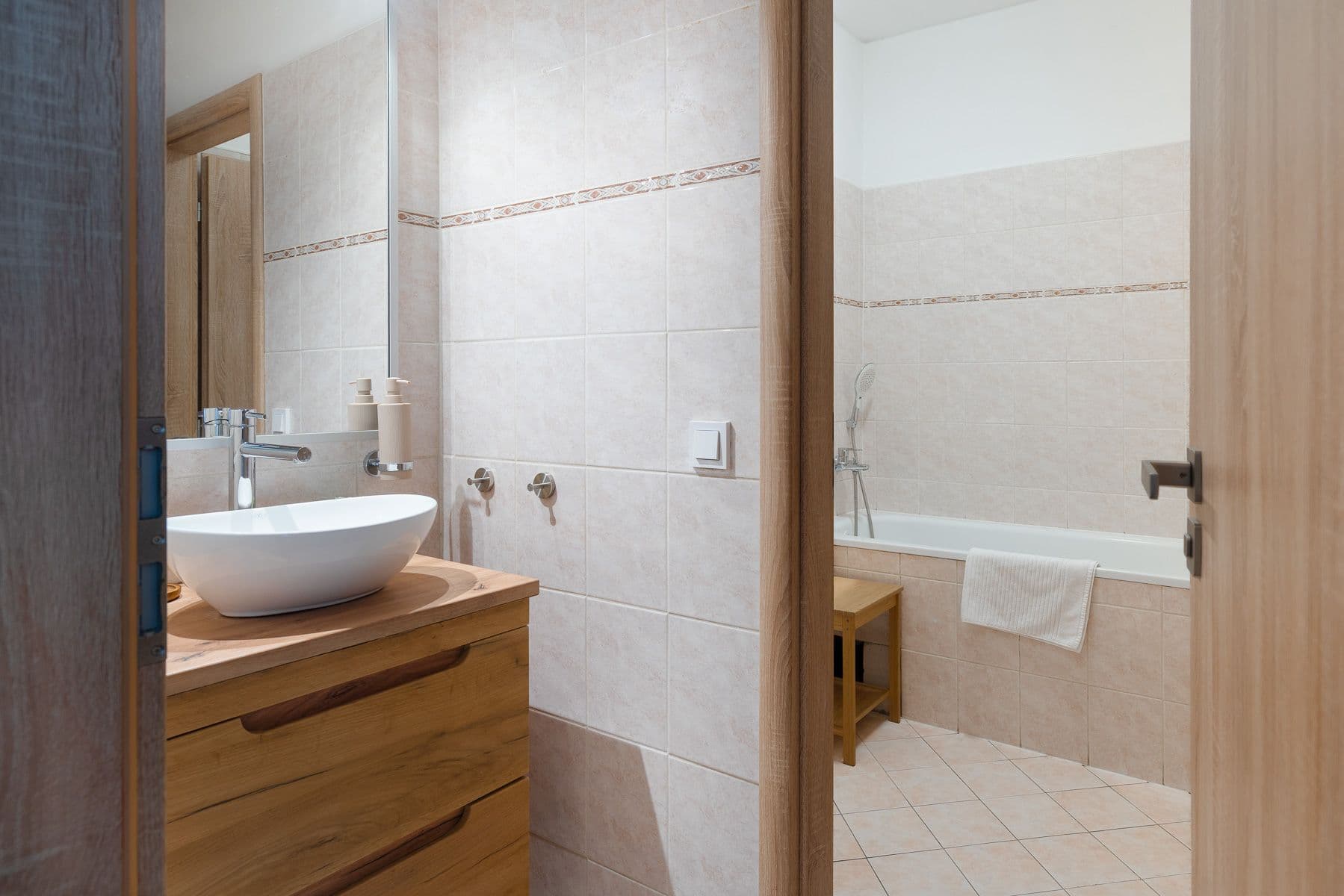 Bathroom with a vessel sink on an oak vanity and beige tiles, with a bathtub and bamboo stool visible through the doorway