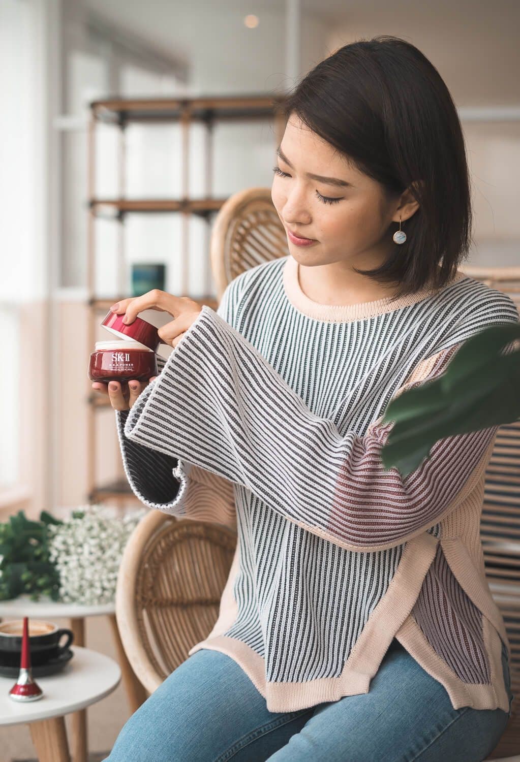 Bella Kuan in a rattan chair opening a jar of SK-II R.N.A. Power cream, coffee and a facial tool beside her.