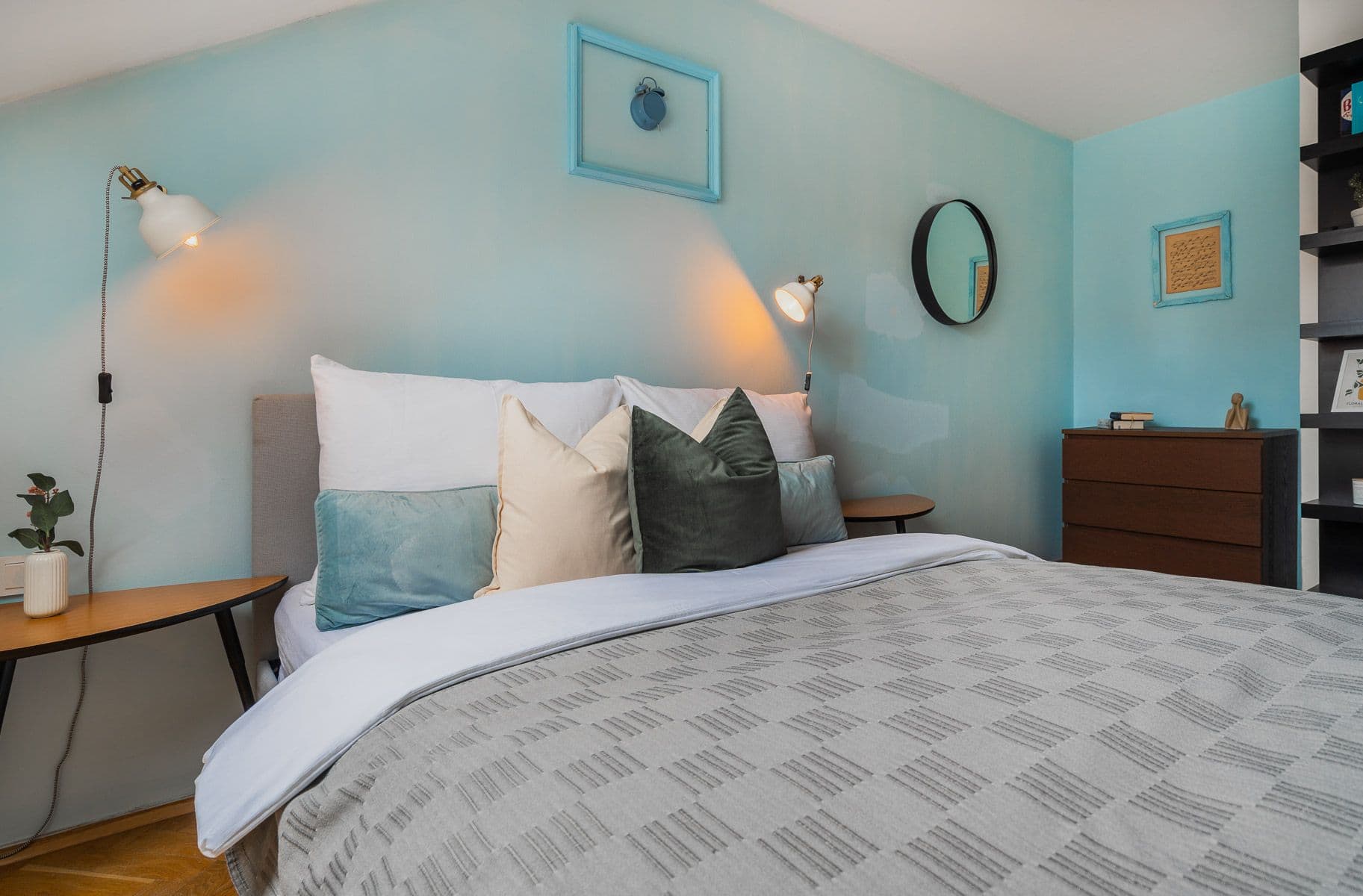 Bed with teal and cream cushions, white wall-mounted reading lamps and a light blue accent wall under a sloped ceiling