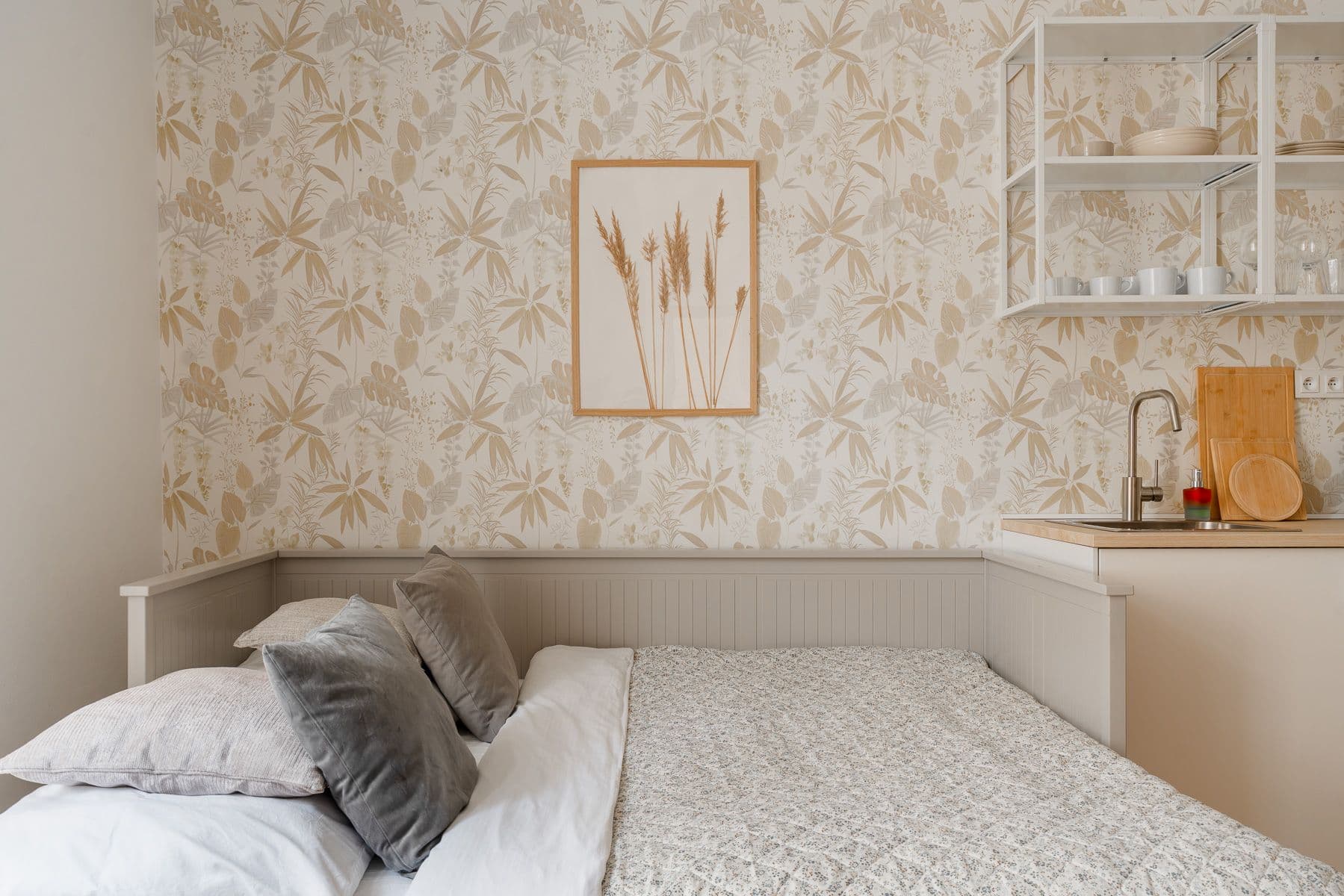 Daybed with grey cushions and a floral quilt against beige botanical wallpaper, next to a framed dried-grass print and open kitchen shelves