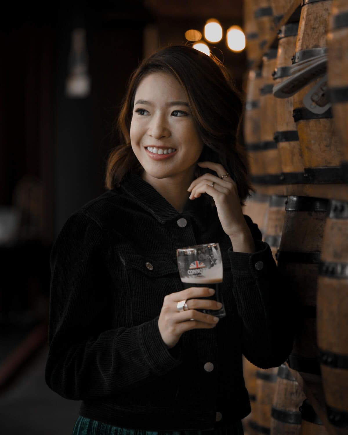 Bella Kuan in a dark corduroy jacket holding a glass of Connor’s stout beside a wall of wooden barrels.