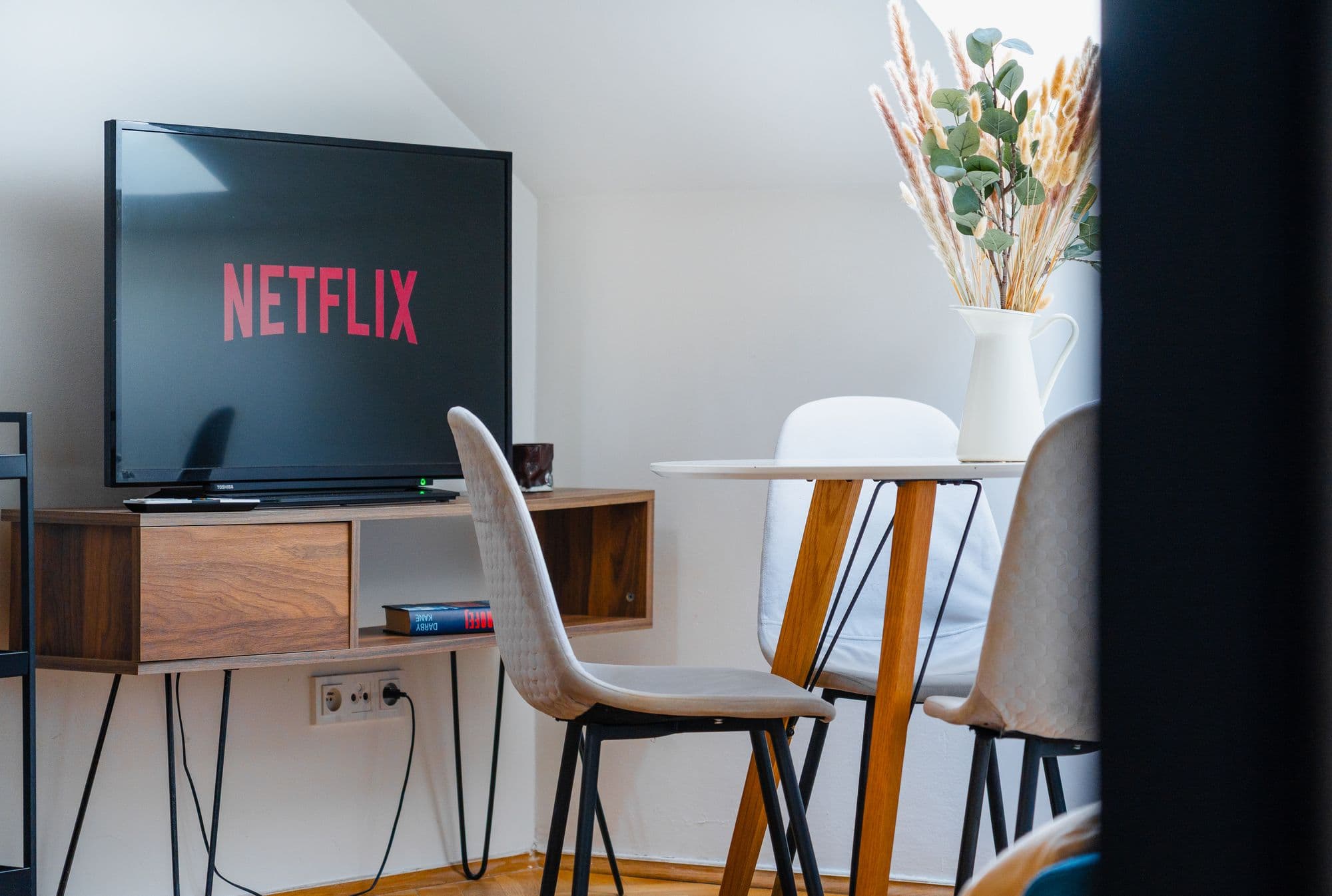 TV on a walnut hairpin-leg console beside a round dining table with a white jug of dried grasses and eucalyptus