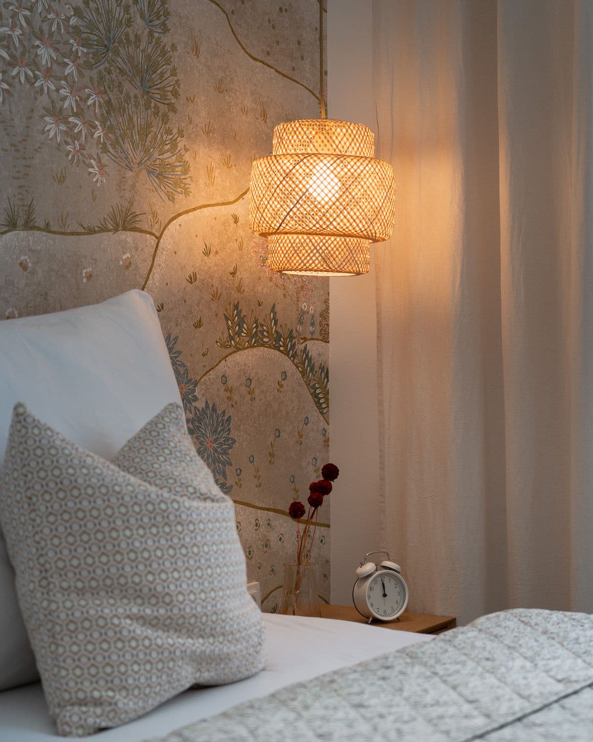 Glowing rattan pendant lamp above a bedside table with dried flowers and a vintage alarm clock