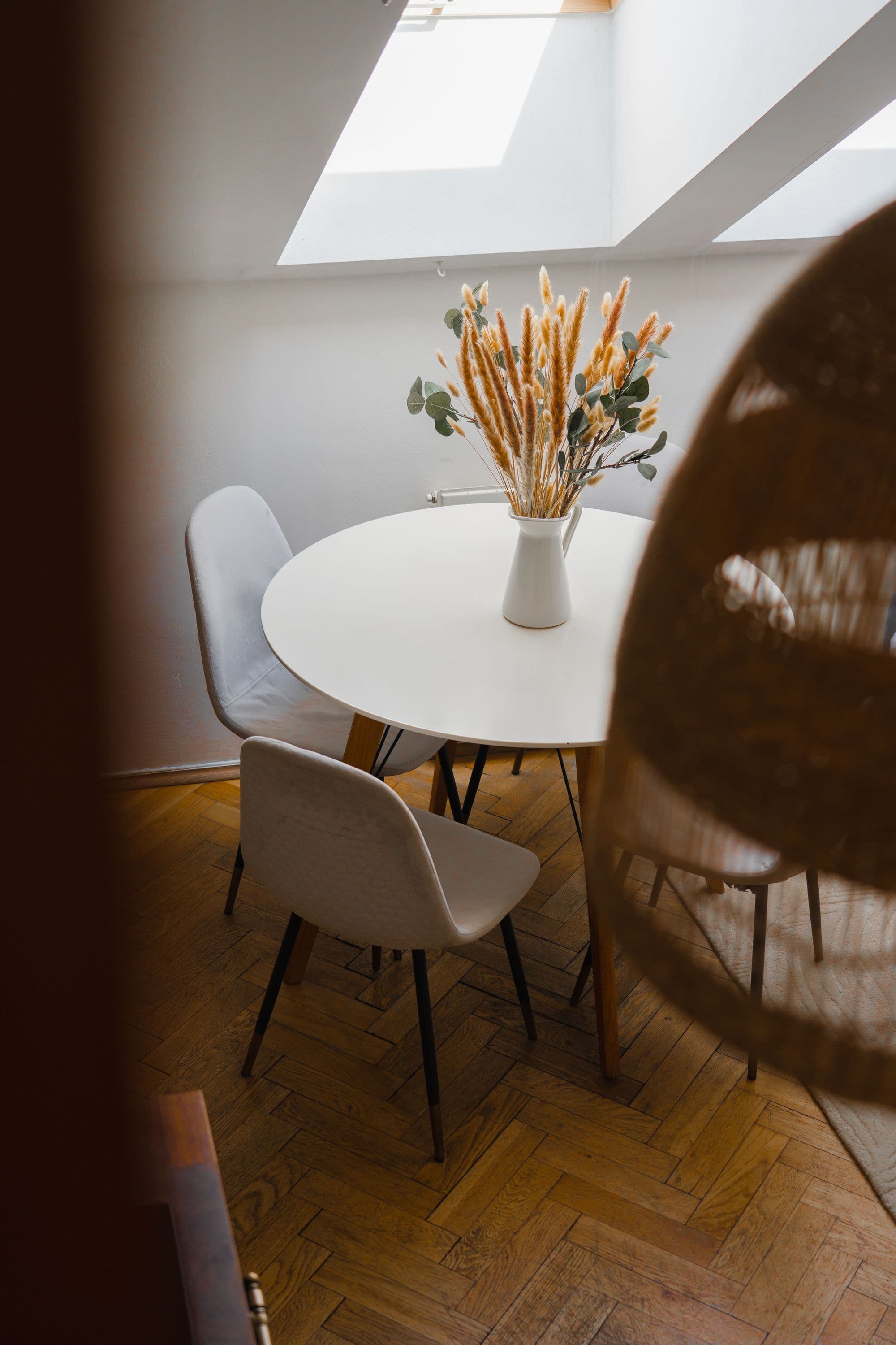 Round white dining table with a bouquet of dried grasses, lit by a skylight above herringbone parquet