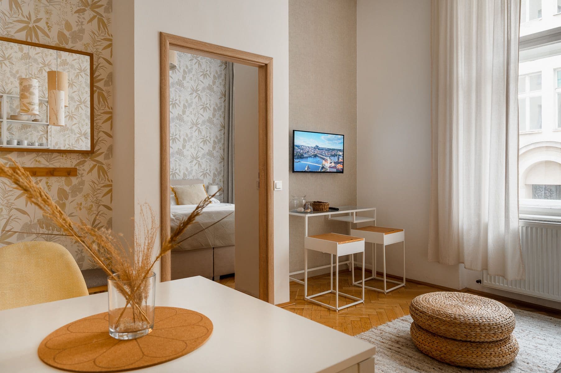 Living room with dried grasses on a dining table, woven poufs, a wall-mounted TV and a doorway into the bedroom