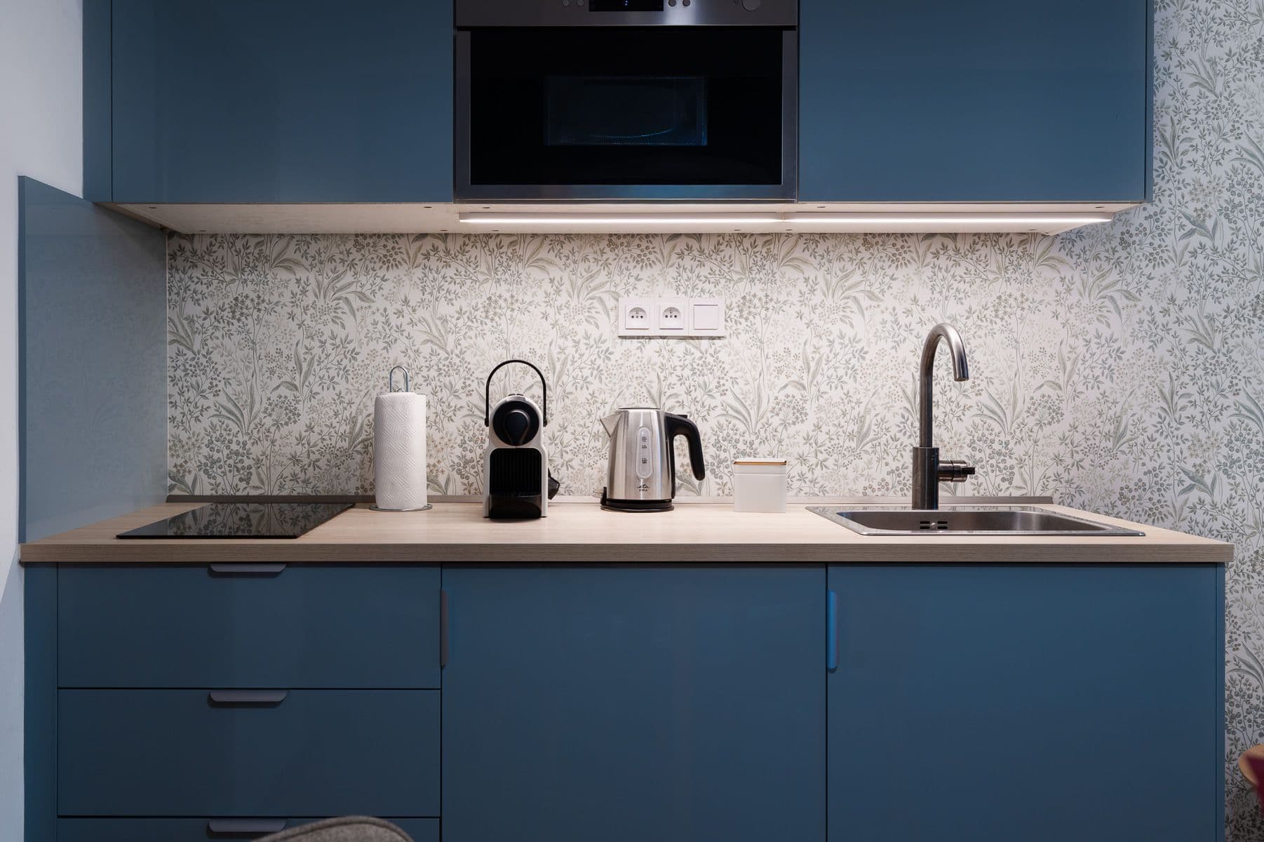 Blue kitchenette with under-cabinet LED lighting, a botanical wallpaper backsplash, coffee machine, kettle and sink