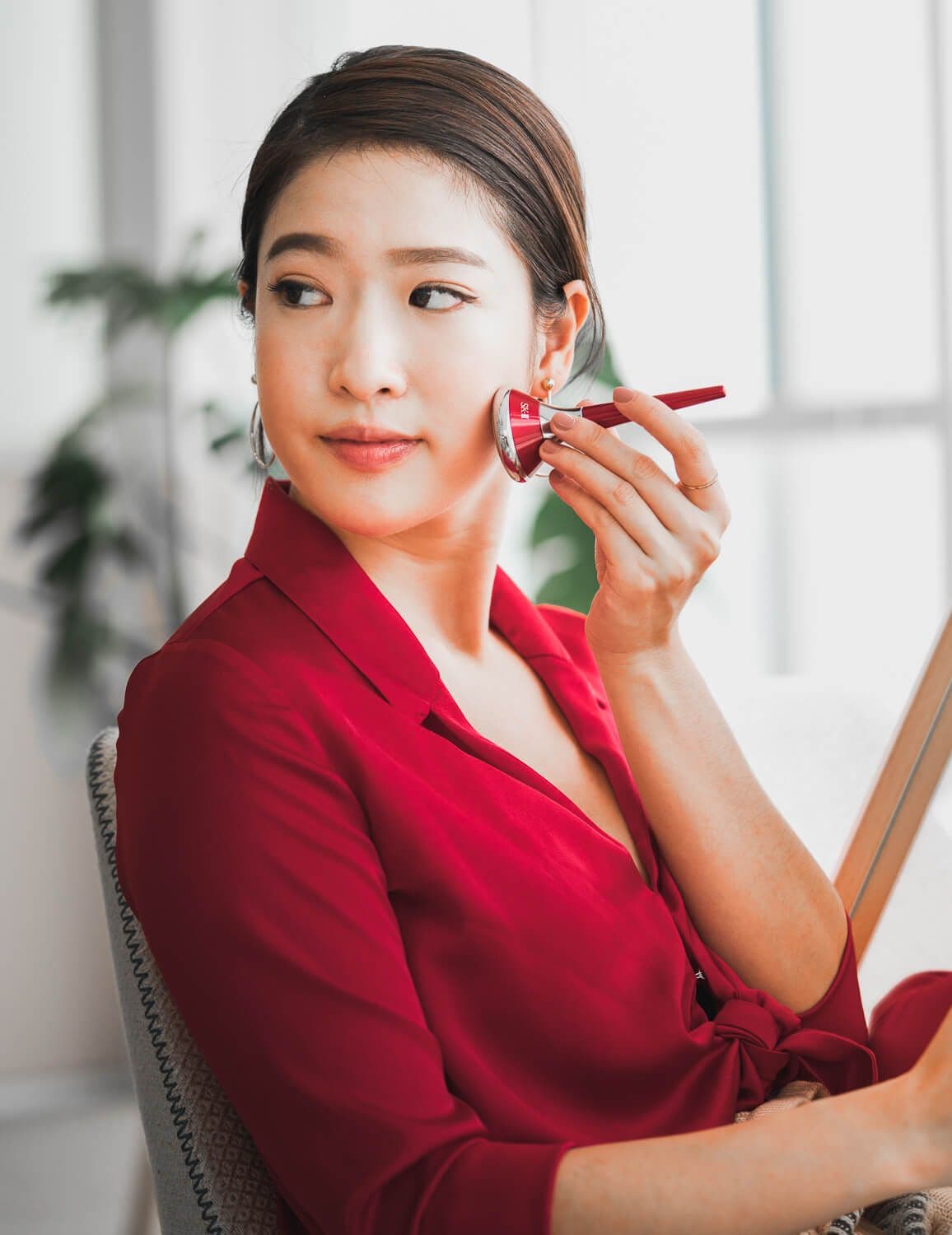 Bella Kuan in a red blouse using an SK-II facial massage tool by a bright window.