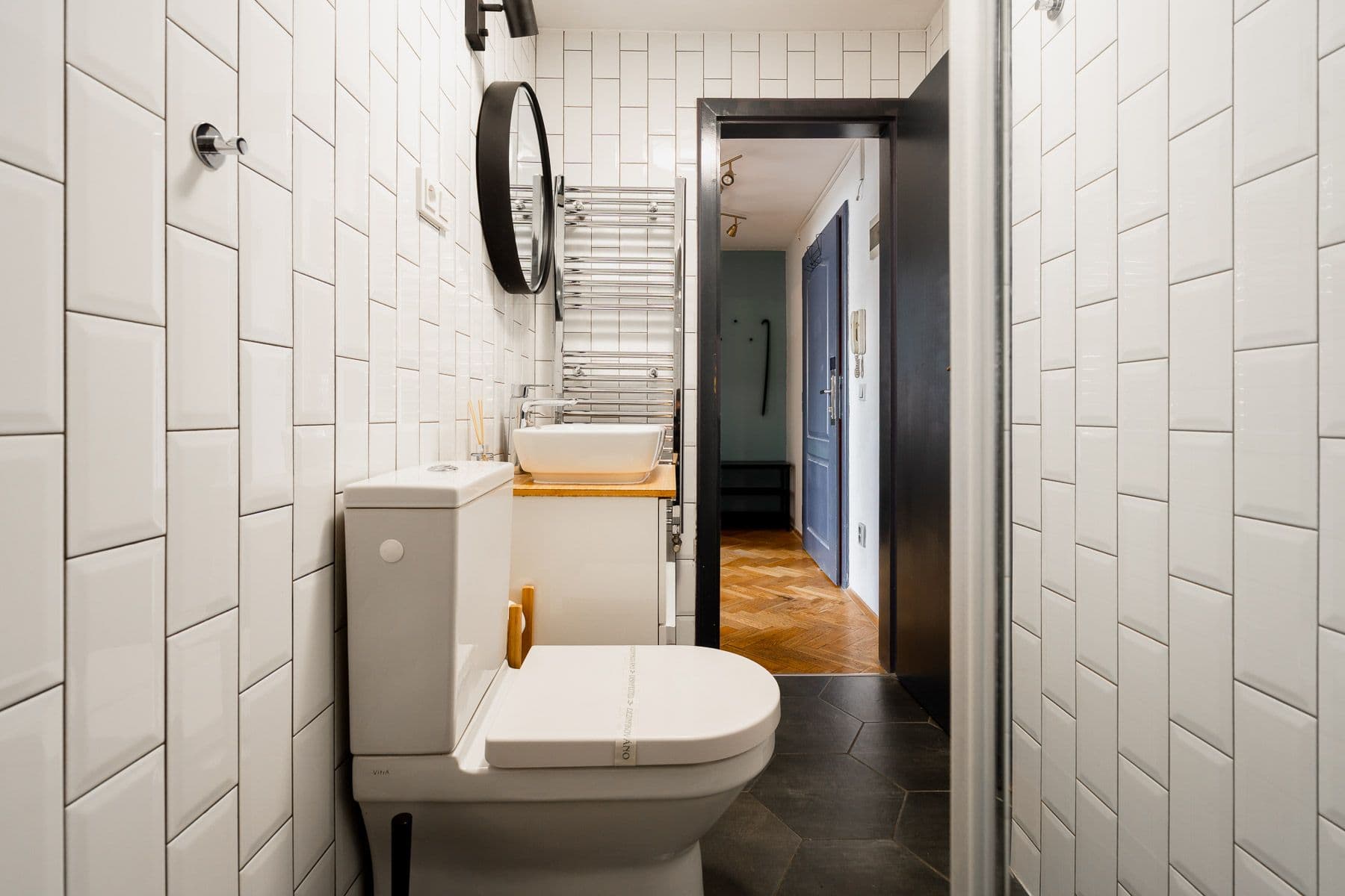 Compact white-tiled bathroom with a round black mirror and chrome towel radiator, door open to a herringbone hallway