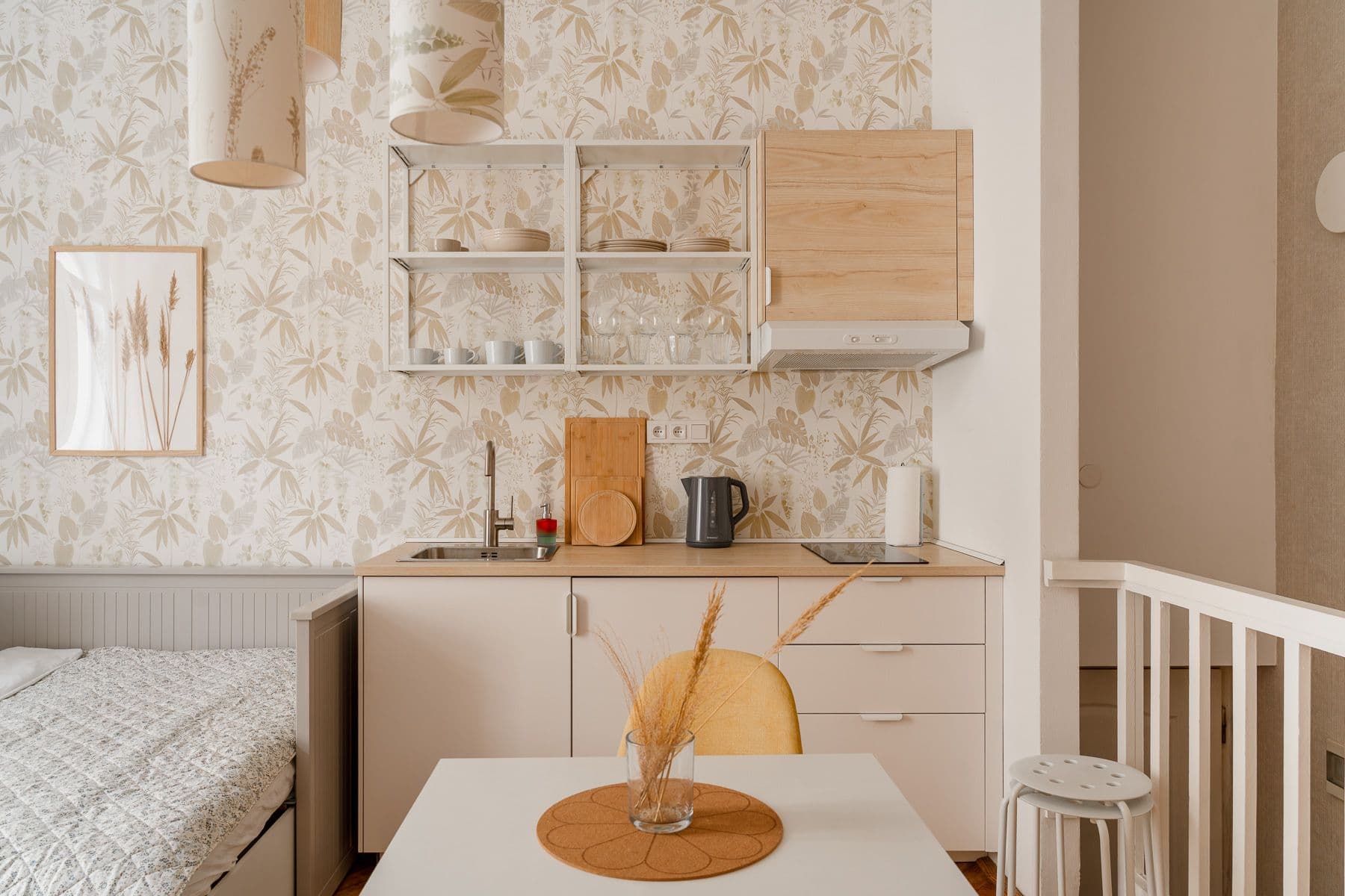 Cream kitchenette with open white shelving, a wood-look wall cabinet, kettle and hob under pendant lamps with botanical shades